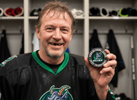 Smiling hockey player in a dark green jersey holding a custom team puck in a locker room